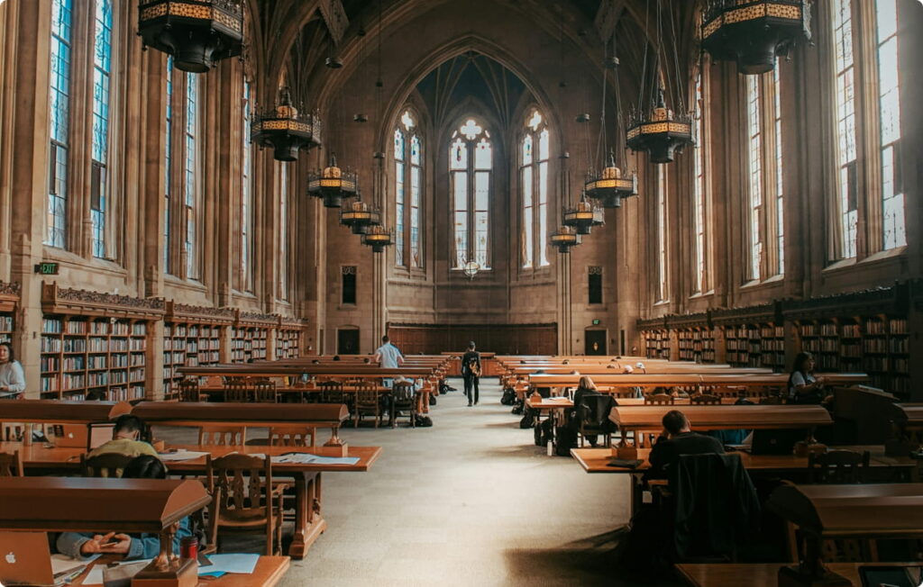 Students studying in a grand library with high vaulted ceilings, hanging chandeliers, and tall arched windows